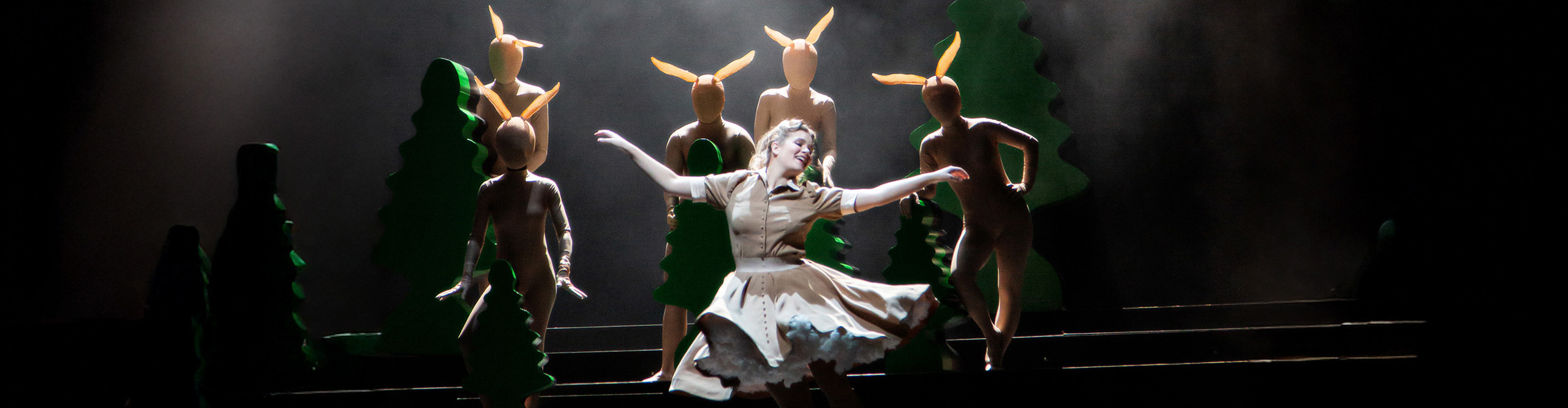 Eine Frau in weißem Kleid steht vor verkleideten Figuren auf einer Bühne.