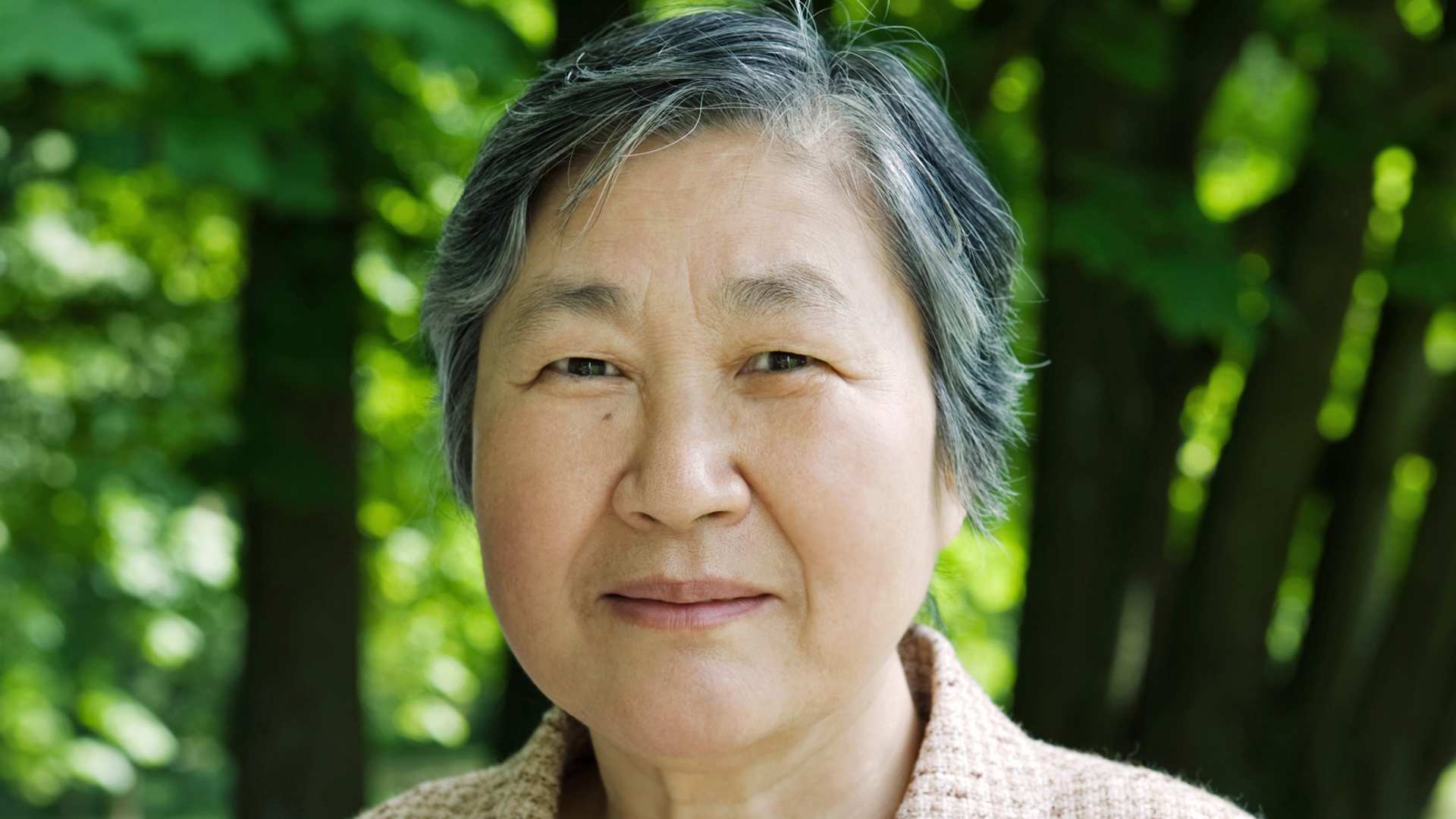 Younghi Pagh-Paan steht in einem hellen Wollpullover vor einem grünen Wald