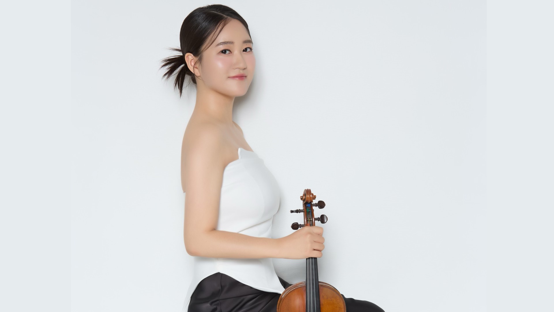 You Bin Min sitzt in einer schwarzen Hose und einem weißen Top auf einem Stuhl und hält eine Geige in der Hand