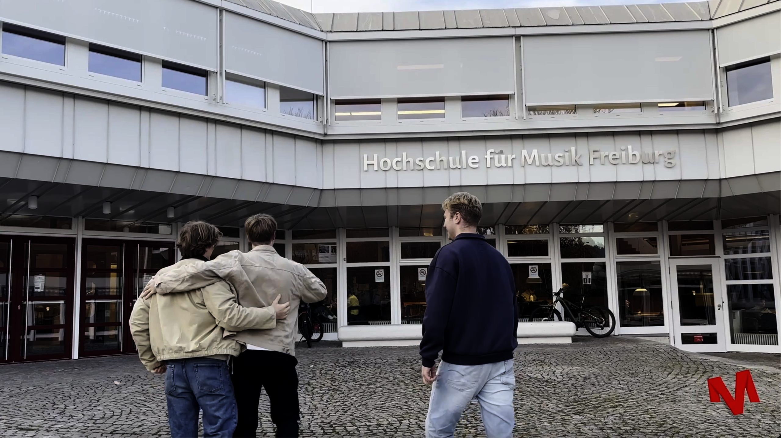 Auf dem Bild sieht man drei Personen von hinten. Eine Person legt den Arm um die andere. Alle drei schauen auf das Hauptgebäude der Hochschule für Musik Freiburg.
