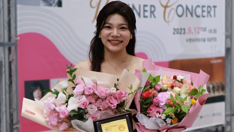 Suji Choi steht mit Blumen und einem Preis in der Hand vor einem Plakat