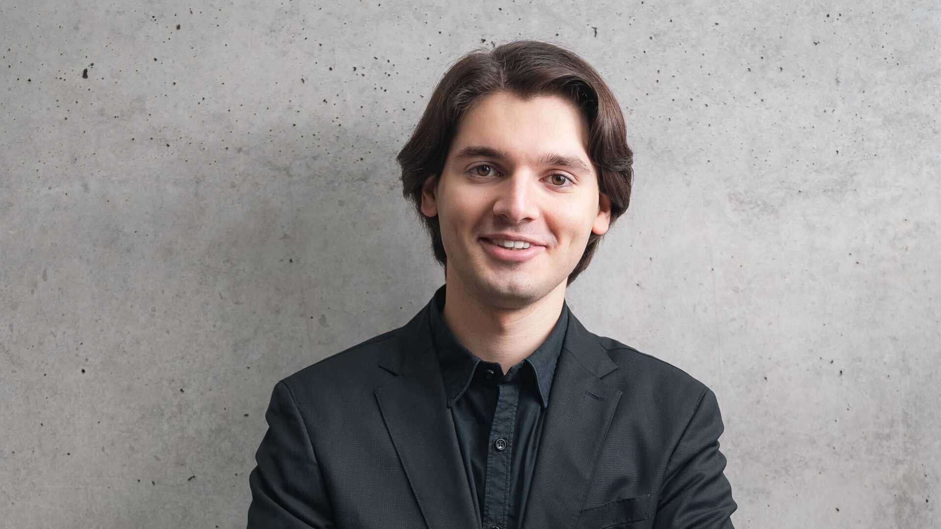 Luis Benedict Alfsmann steht in einem schwarzen Anzug vor einer grauen Wand