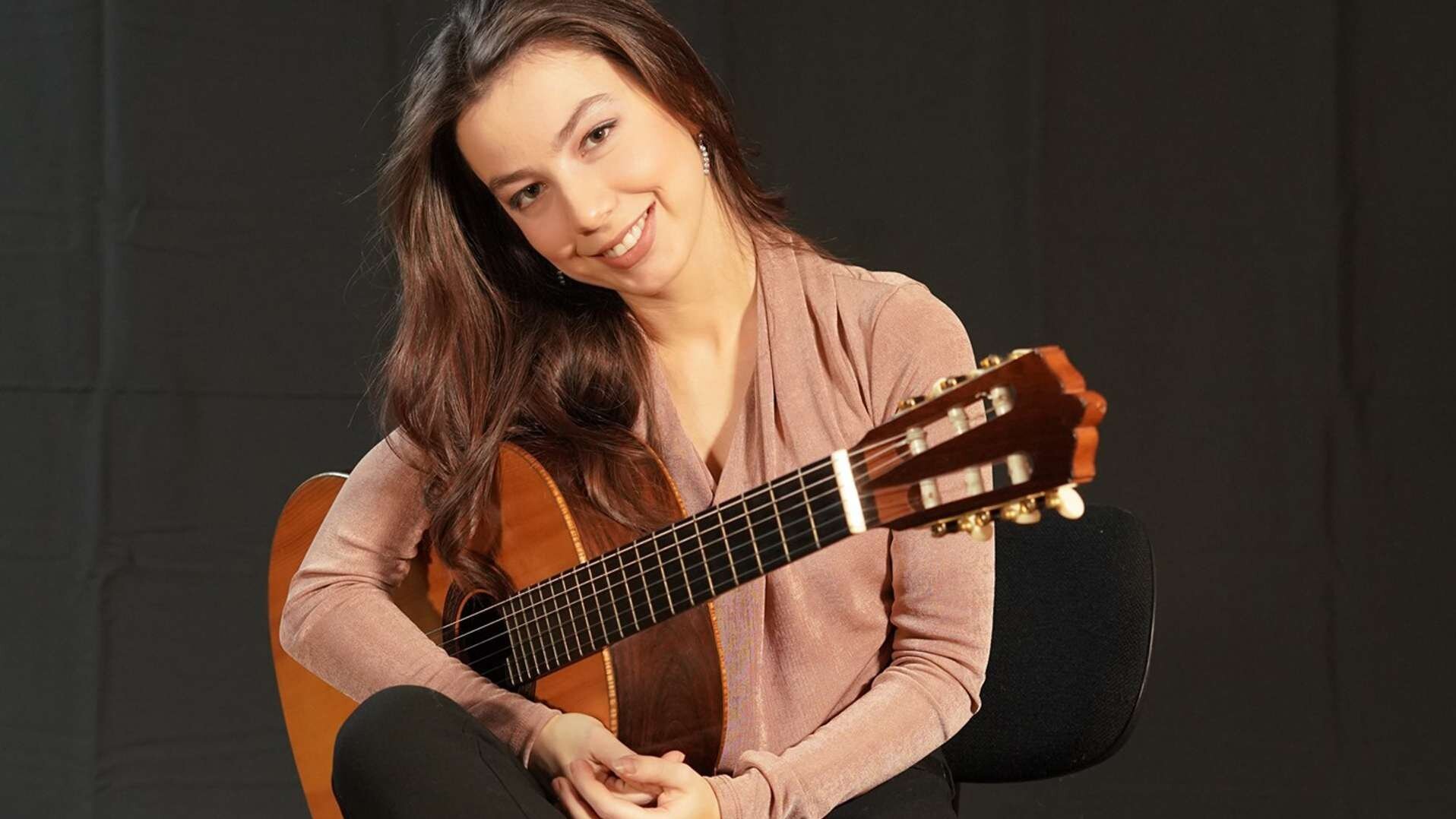 Nisa Metlillo sitzt mit Gitarre unter dem Arm auf einem Stuhl