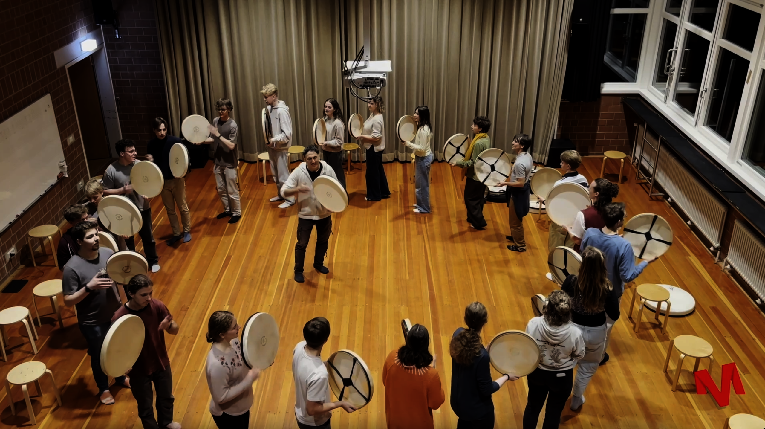 Man sieht eine Gruppe von Menschen die in einem Kreis mit Trommeln musizieren. In der Mitte steht eine andere Person die auch eine Trommel in der Hand hält.