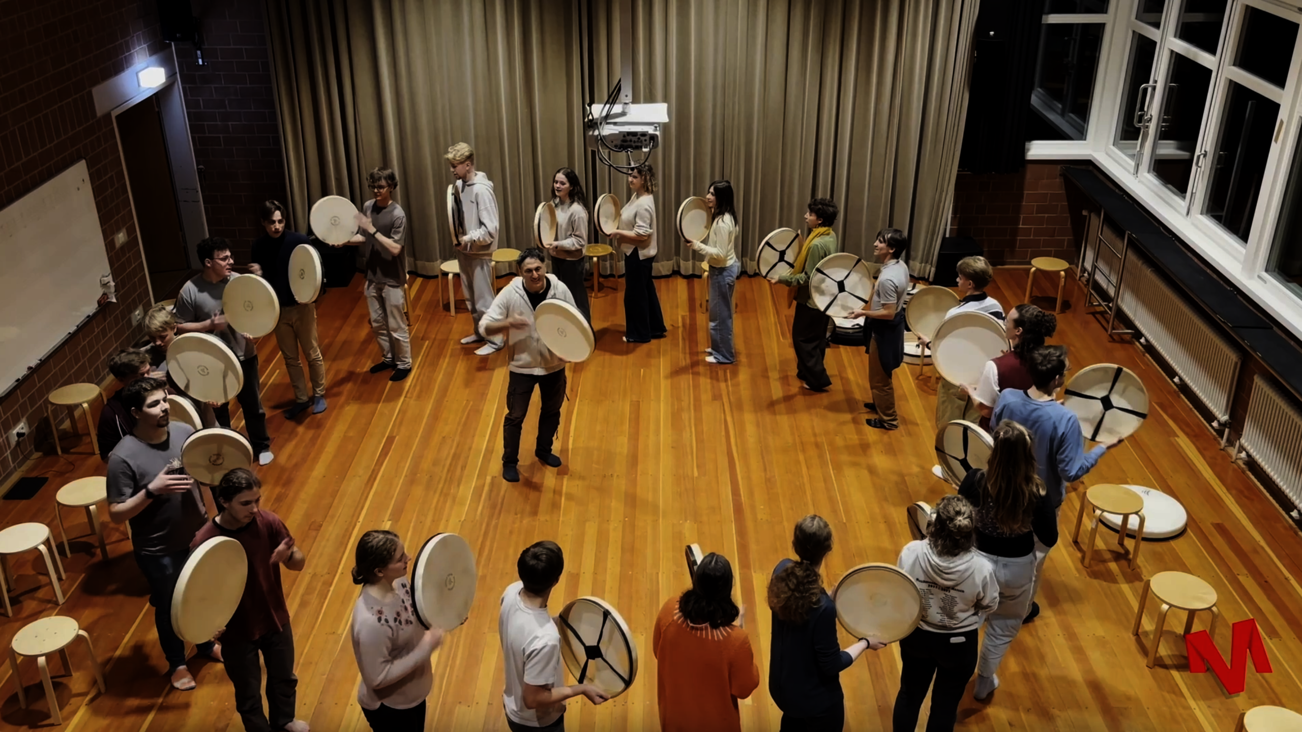Man sieht eine Gruppe von Menschen die in einem Kreis mit Trommeln musizieren. In der Mitte steht eine andere Person die auch eine Trommel in der Hand hält.