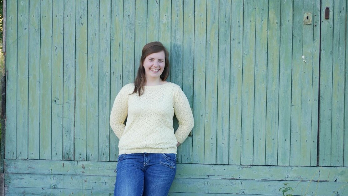 Annika lehnt an einer Wand. Die Arme sind hinter dem Rücken verschränkt.