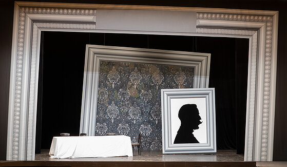 Drei riesige, weiß-silberne Bilderrahmen umrahmen die Bühne der Hochschule, im Vordergrund stehen ein langer Tisch und drei Stühle