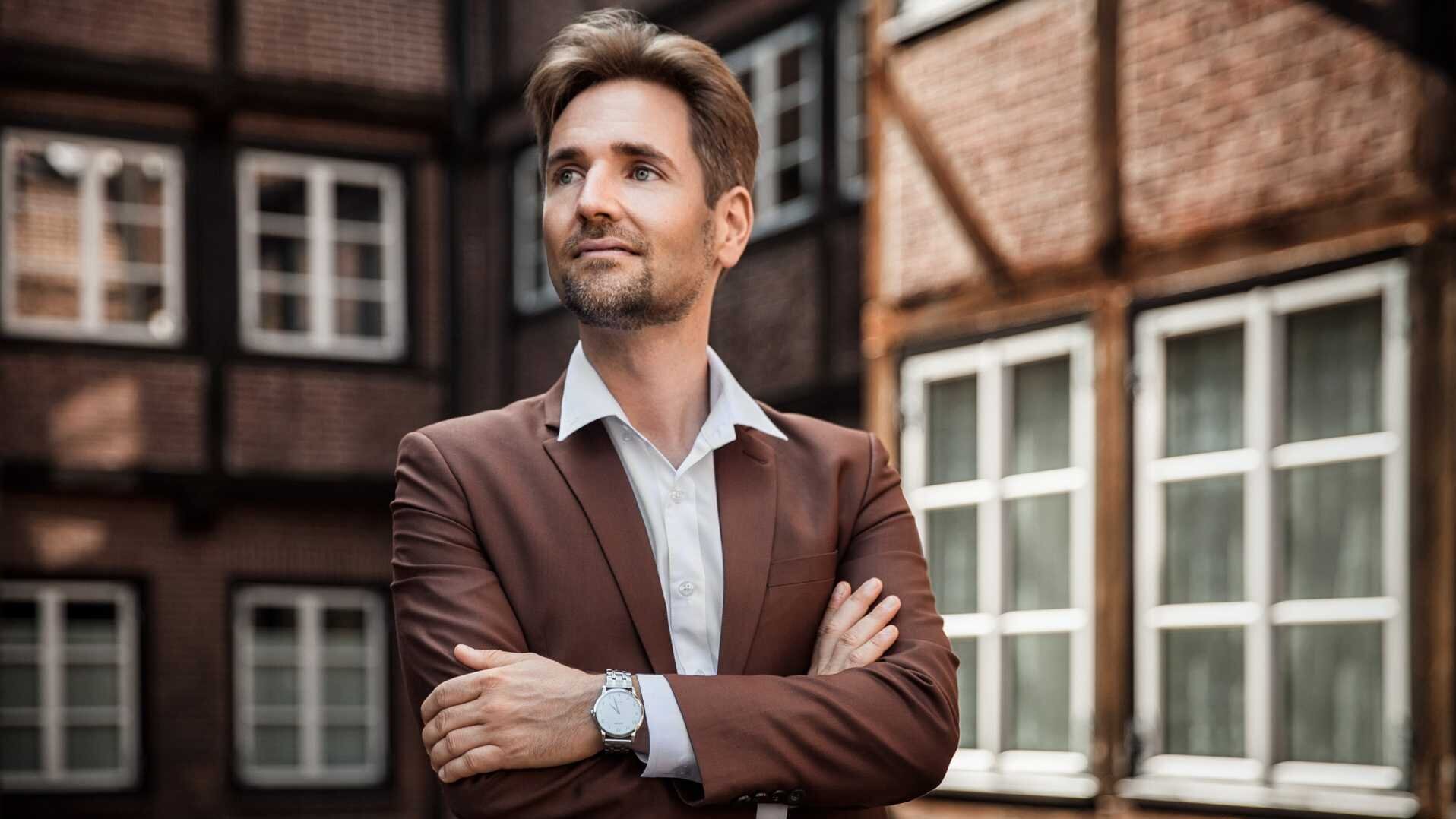 Laurens Patzlaff steht in einem braunen Anzug vor einem Fachwerk-Haus